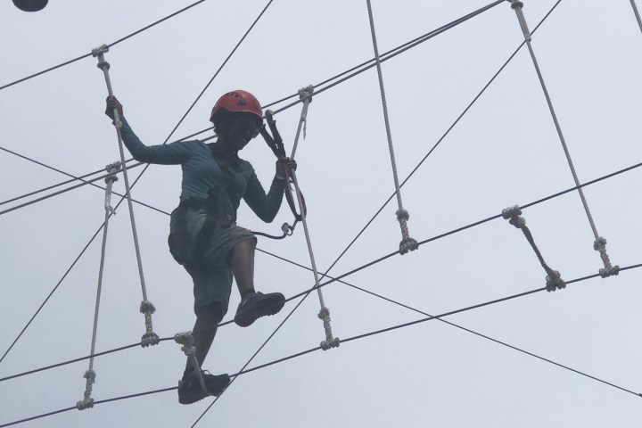 Person in helmet crossing a high ropes course with a cloudy sky background.
