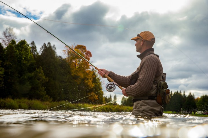 Person fly fishing in a river, surrounded by trees and cloudy sky.