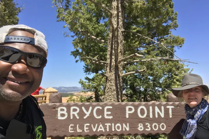Two people posing by a Bryce Point sign under a tree, clear sky in the background.