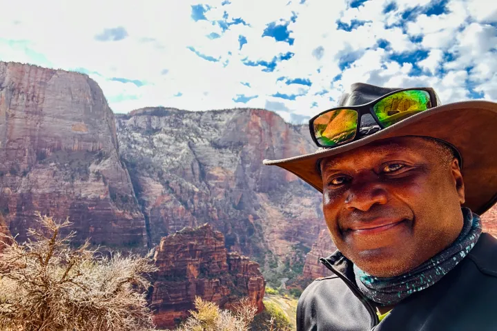 Man in hat with sunglasses on rim, rocky canyon background, blue sky with clouds.