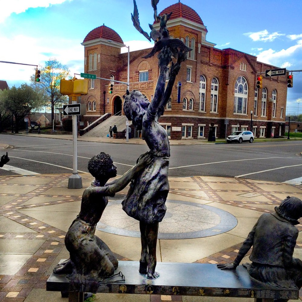 Bronze statues of children playing near a historic brick building on a city street corner.