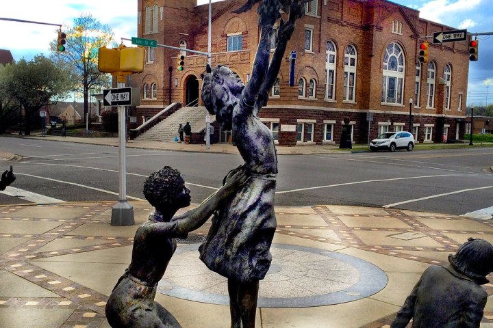 Bronze statues of children playing near a historic brick building on a city street corner.