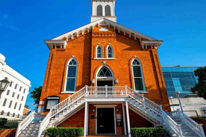 Historic red-brick church with white stairs and arched windows under a blue sky.