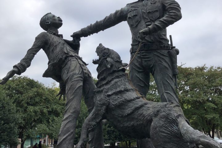 Bronze statue of a police officer, dog, and civilian on a rock platform under a cloudy sky.