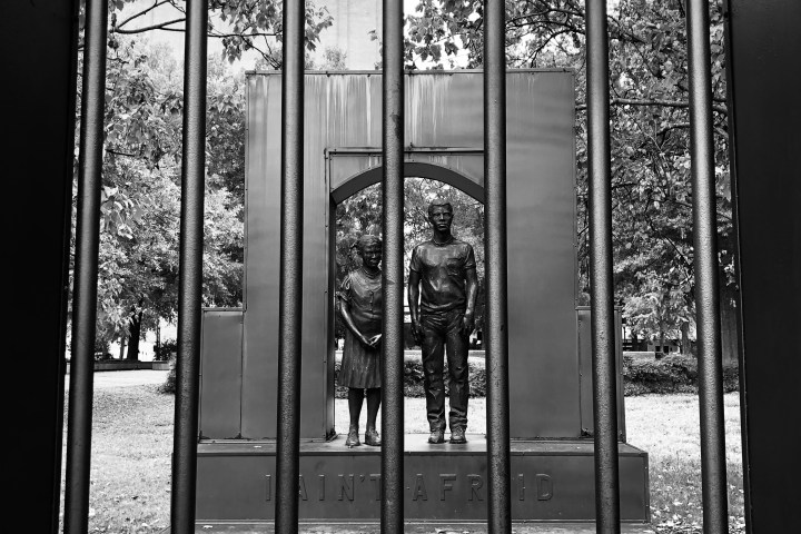 Black and white photo of statues seen through vertical metal bars, with trees in the background.