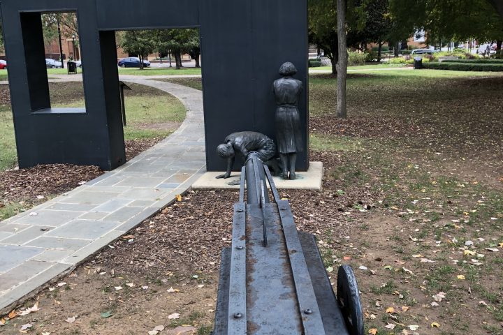 Sculpture of a large gun aimed at two figures near a park walkway.