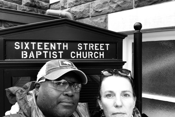 Two people in front of Sixteenth Street Baptist Church sign; one wears a cap, the other sunglasses on head.