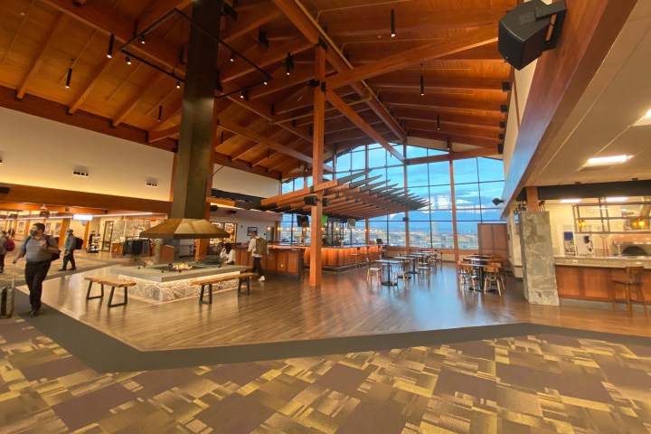 Spacious airport lounge with wooden beams, large windows, tables, and a central fireplace.