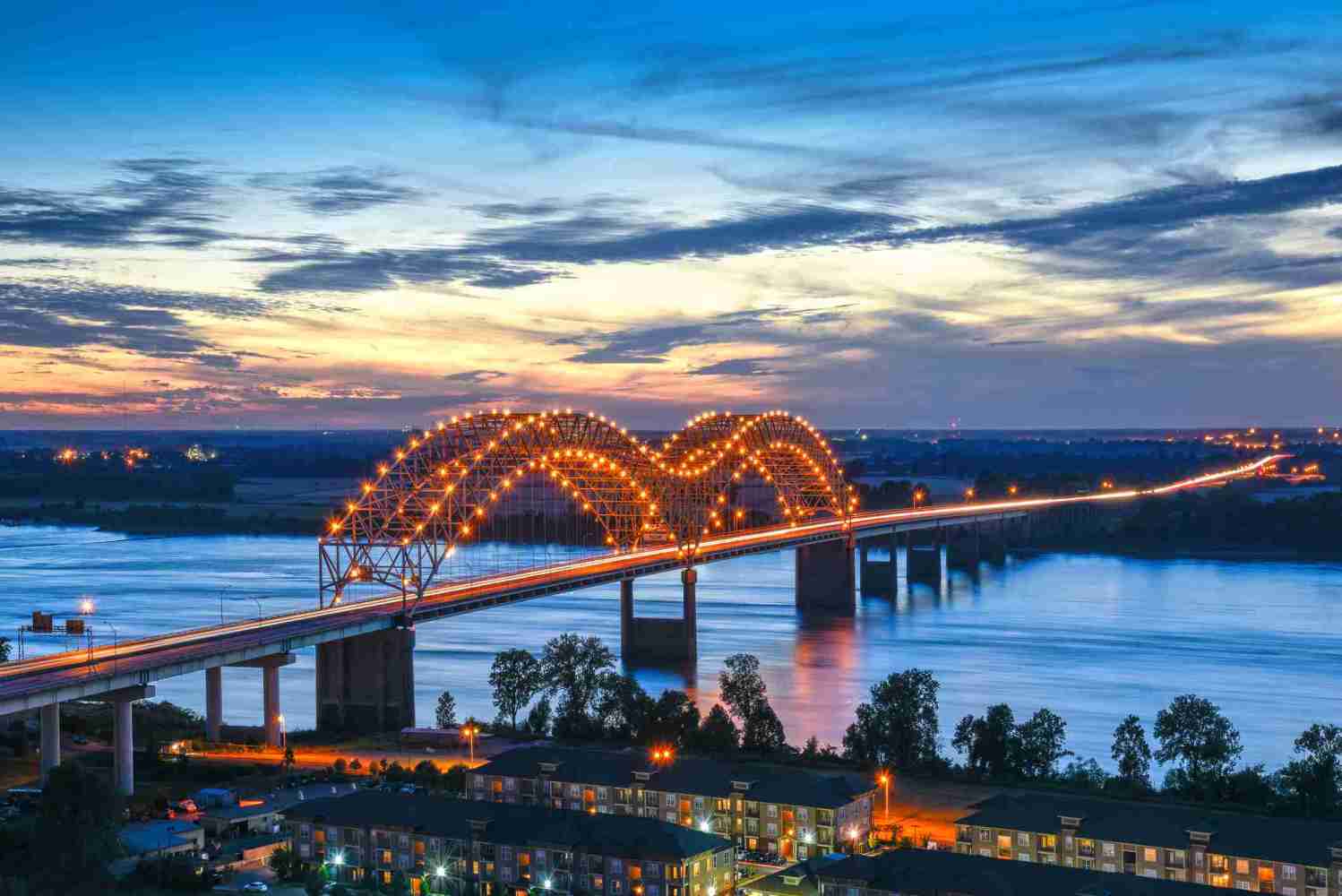 Illuminated bridge over river at dusk with colorful sky and city lights in background.