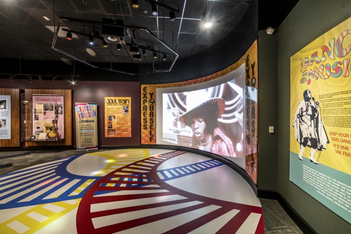 Colorful museum exhibit with music memorabilia, posters, and a large TV screen showing a performer.
