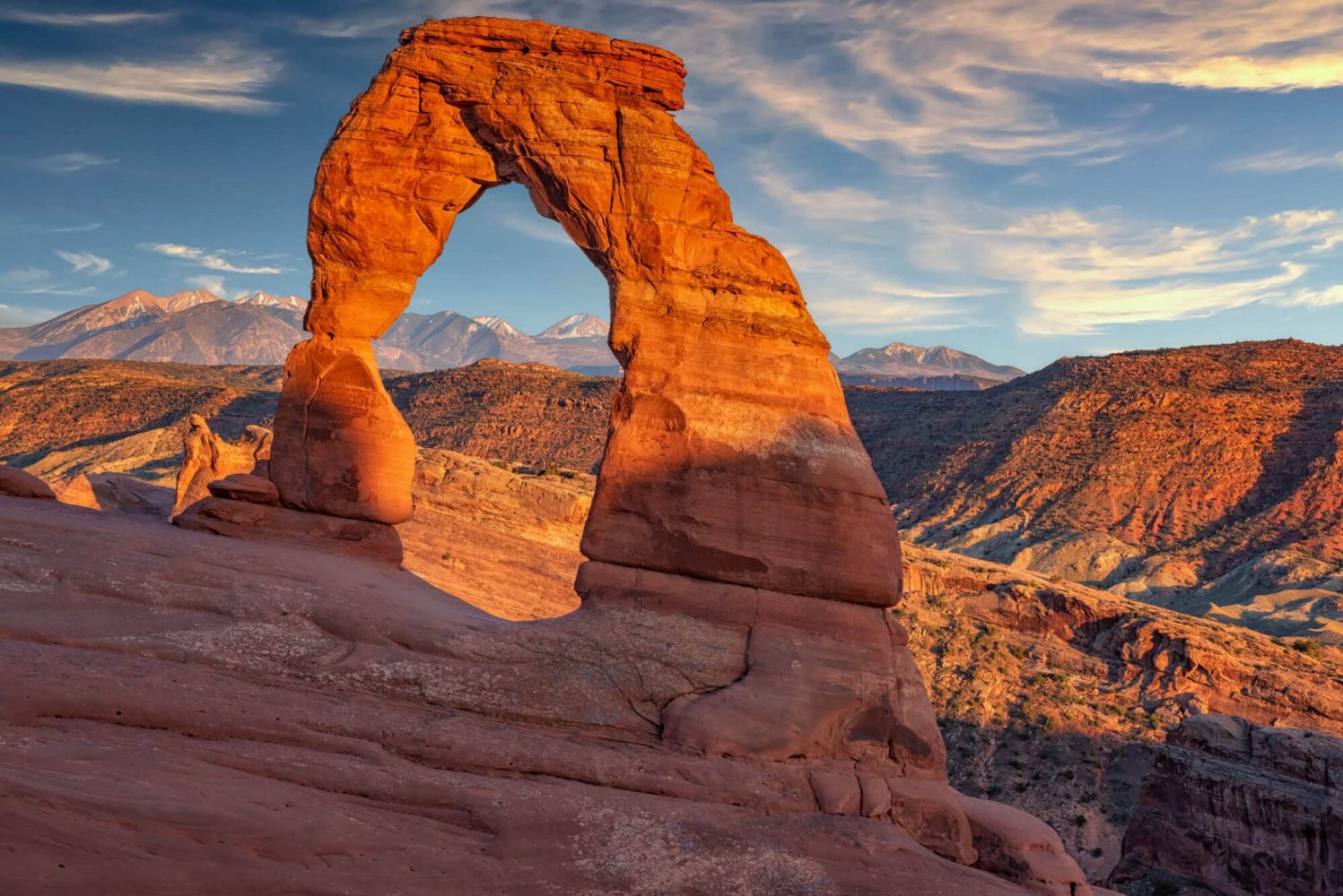 Delicate Arch at sunset with mountains and blue sky in the background.