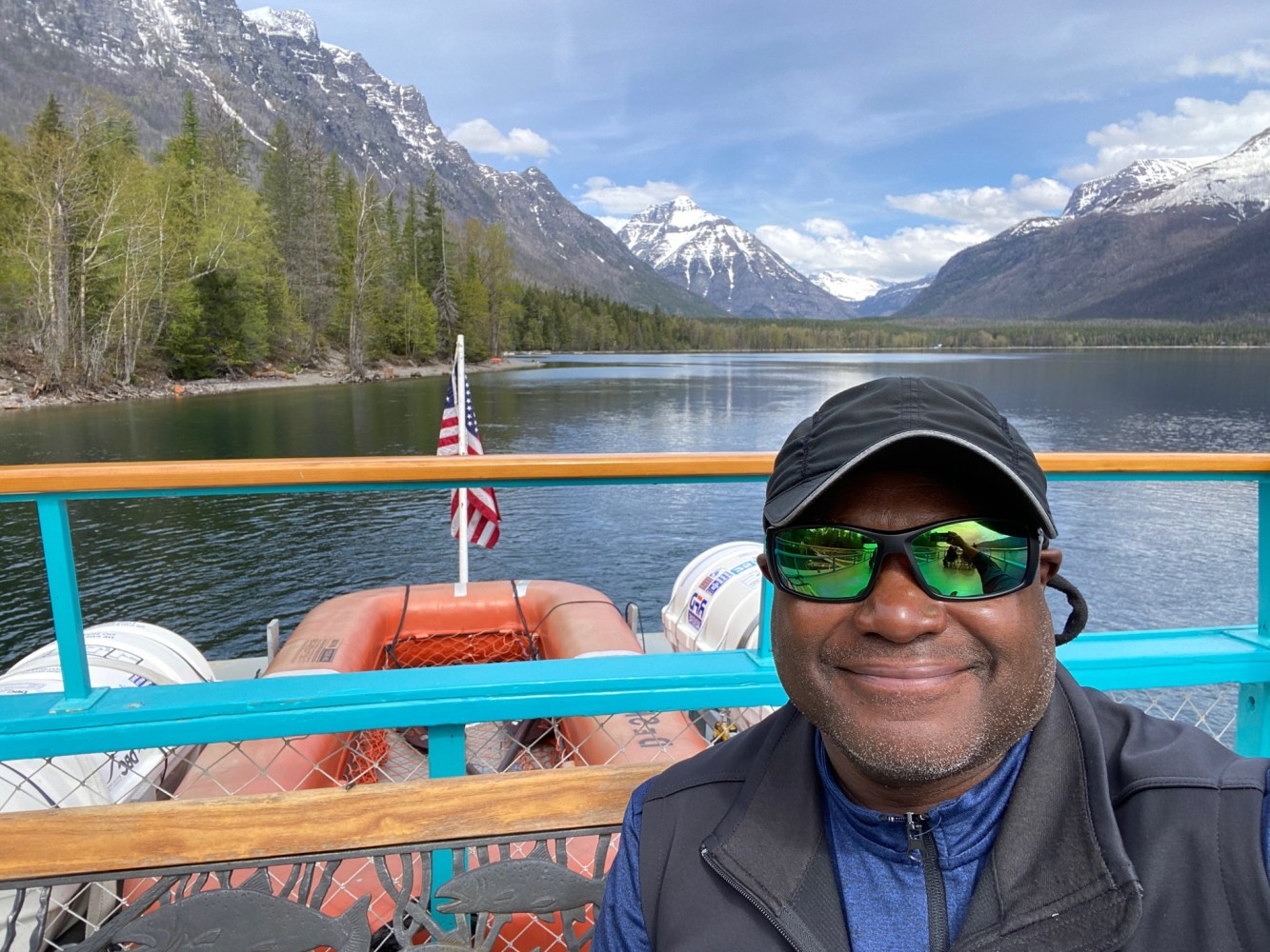 Smiling man in sunglasses on a boat with mountains and lake in the background.