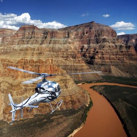 Helicopter flying over a canyon with a brown river in sunlight.
